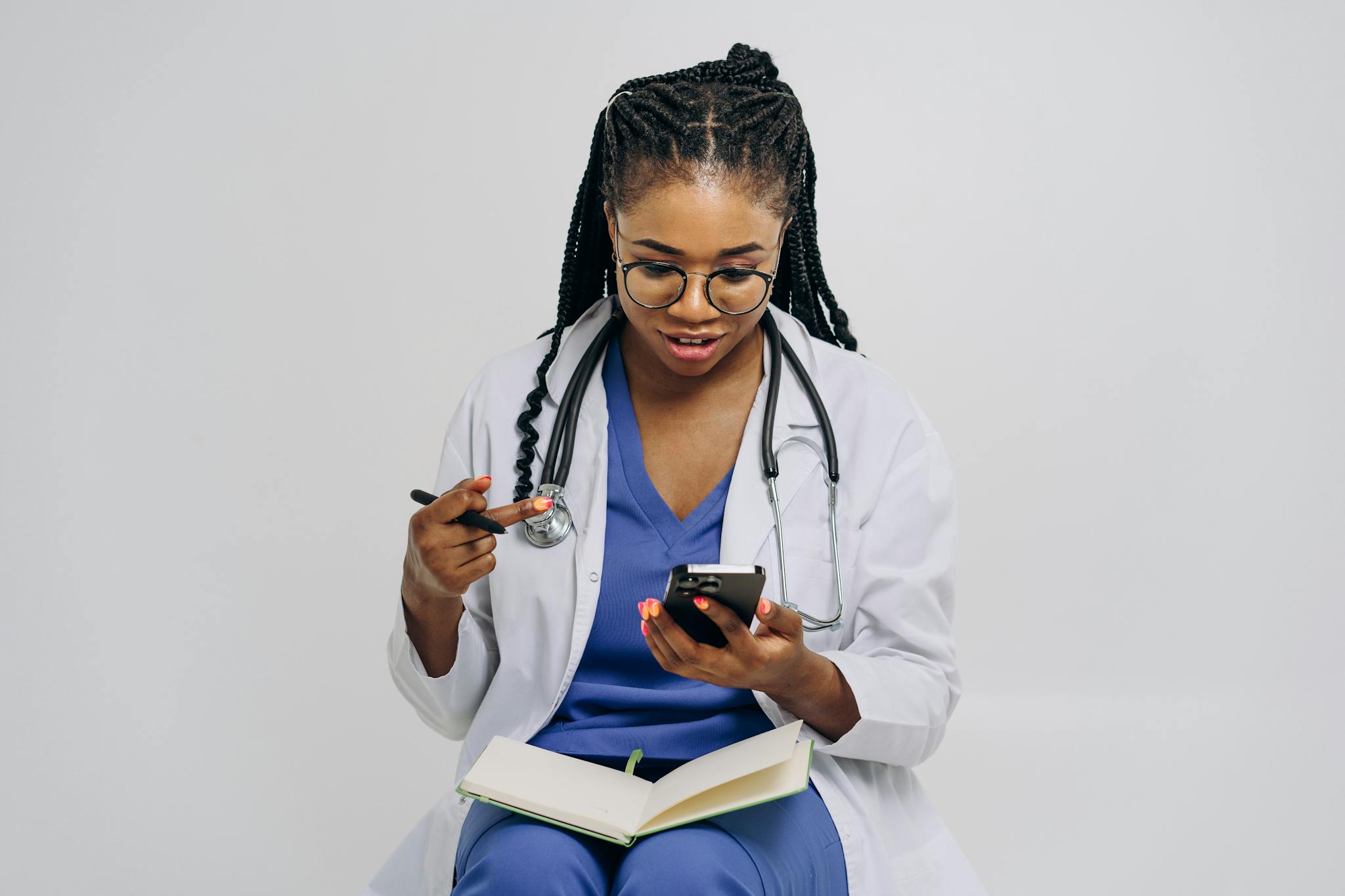 Black female doctor checks smartphone while holding notebook on white background.